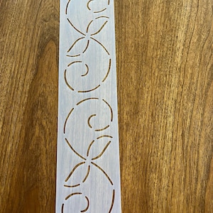 May include: A rectangular stencil with a repeating floral design. The stencil is white and has a small hole at the top for hanging. The floral pattern consists of curved lines and petal shapes, creating a decorative element.
