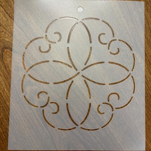 May include: A stencil with a floral design cut out. The design features curved lines and petal-like shapes arranged in a circular pattern. The stencil is made of a translucent, light-colored material and has a small hole at the top for hanging.