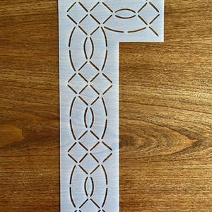 May include: White plastic quilting stencil with a design of interlocking circles and diamonds. The stencil is shaped like a capital letter "L".