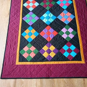 May include: A quilted blanket featuring a central black panel with a grid of multi-colored square patches in shades of blue, green, purple, orange, and red. The quilt is framed by a thin yellow border and a wide, quilted maroon outer border.