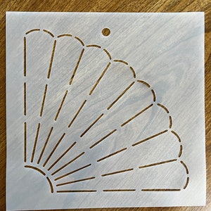 May include: A white stencil featuring a fan or sunburst design. The design is cut out, revealing the surface beneath. The stencil has a small hole at the top for hanging. Ideal for crafts and DIY projects.