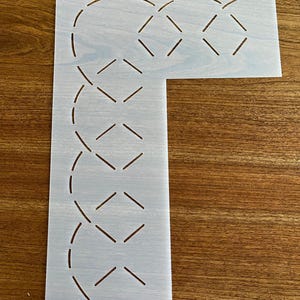 May include: A light-colored stencil with a repeating geometric pattern of curved lines and diamond shapes. The stencil is rectangular with a right-angle cut-out. A small hole is at the top for hanging. The stencil is on a wooden surface.