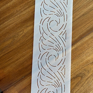 May include: A rectangular stencil with a repeating pattern of curved lines and swirls. The stencil is white with a series of cut-out shapes. The design is symmetrical and features a series of curved lines and swirls. The stencil has a small hole at the top for hanging.
