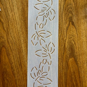 May include: A rectangular stencil with a floral design. The stencil is light gray with a series of leaf-like cutouts arranged in a vertical pattern. A small, round sticker with the number "#30" is attached to the top.