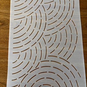 May include: A white stencil with a repeating pattern of curved lines. The lines are cut out of the stencil, creating a negative space design. The stencil is rectangular and has a hole at the top.