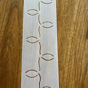 May include: White plastic stencil with a repeating pattern of oval shapes and wavy lines. The stencil is designed for quilting or other crafts.