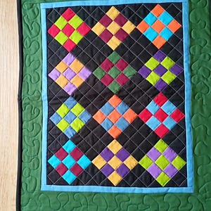 May include: A rectangular quilted wall hanging featuring a black background with a grid of multi-colored squares in shades of red, lime green, yellow, purple, orange, and blue. The quilt has a light blue inner border and a wide green outer border.