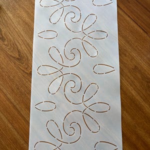 May include: A rectangular, semi-transparent plastic stencil featuring a repeating pattern of interconnected, dashed-line swirls and teardrop shapes. The stencil has a single small circular hole at the top center for hanging or alignment purposes.