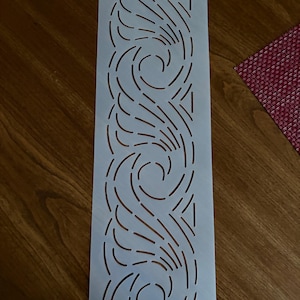 May include: A rectangular white plastic stencil featuring a repeating pattern of curved, swirling lines. The design consists of elongated, segmented cutouts that create a flowing, wave-like motif. A small circular hole is positioned at the top center.
