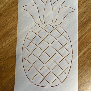 May include: A white plastic stencil of a pineapple with a stitched design. The pineapple is outlined with a dashed line and the leaves are detailed with a stitched pattern.