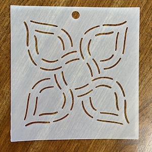 May include: A square, off-white stencil with a floral design cut out. The design features four curved petal shapes, each with curved and straight lines. A small hole is at the top for hanging. The stencil is on a wooden surface.