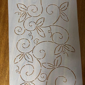May include: A stencil with a floral design featuring swirling lines and leaf shapes. The stencil is made of a light-colored, semi-transparent material and has a small hole at the top for hanging. The design is cut out, creating negative space.