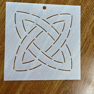 May include: White plastic quilting template with a geometric design of intersecting curved lines. The template has a small hole in the top center.