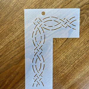May include: A white plastic stencil with a decorative, intricate design of a braided rope pattern. The stencil is shaped like a right angle, with a longer vertical side and a shorter horizontal side.