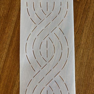 May include: A rectangular, off-white stencil with a repeating, interwoven design. The design features curved and straight lines, creating a braided pattern. A small hole is at the top for hanging or storage. The stencil is on a wooden surface.