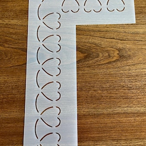 May include: A light-colored, L-shaped stencil with a repeating heart-shaped design. The stencil has a small hole at the top and the number "#21" in a small circle. The stencil is placed on a wooden surface.