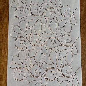 May include: A stencil with a repeating pattern of stylized leaves and swirls. The design is cut out of a translucent, off-white material. The pattern is symmetrical and suitable for various craft projects.