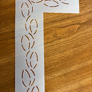 May include: A translucent, L-shaped plastic stencil featuring a repeating pattern of cut-out leaf or petal shapes arranged along the vertical edge. The stencil includes a small circular hole at the top corner for hanging or storage purposes.