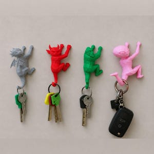 以下が含まれることがあります： グレー、赤、緑、ピンクのクライミングフィギュア型ウォールフック4点。各フィギュアはローポリゴンの幾何学的なデザインで、金属製のキーリングを保持しています。ピンクのフィギュアには黒い車のキーフォブを含む複数の鍵が取り付けられています。