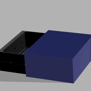 May include: A black rectangular box with a dark blue lid. The box is open, revealing a black interior with circular features. The lid is a solid, dark blue color. The box is on a gray surface.