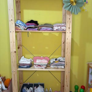 Op de afbeelding: Een houten stellingkast met meerdere planken met netjes opgevouwen kinderkleding. De kleding is in verschillende kleuren, waaronder roze, blauw en wit. De kast staat tegen een gele muur, met decoratieve elementen.