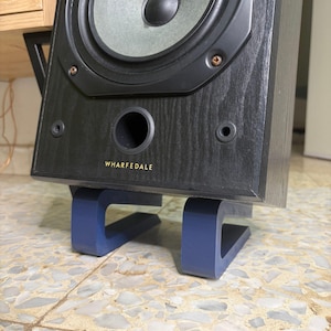 Puede incluir: Un altavoz Wharfedale negro con un cono de altavoz gris, montado sobre un soporte azul. El altavoz tiene un puerto circular y la marca "WHARFEDALE" en letras doradas.