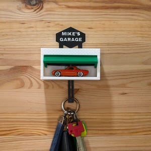 May include: A key holder with a miniature orange car inside a white garage with a green door. The sign above the garage reads "MIKE'S GARAGE" in white text on a black background. A set of keys hangs below the garage.
