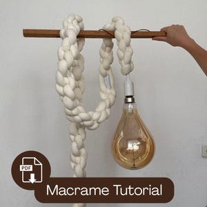 May include: A macrame lamp design featuring a thick, off-white woven cord suspended from a wooden beam. A large, teardrop-shaped light bulb hangs below. The image includes the text "Macrame Tutorial".