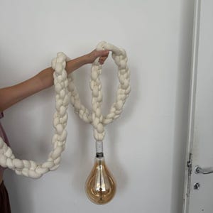 May include: Cream-colored, braided rope light fixture with a large, teardrop-shaped, amber-toned light bulb. The braided rope is held up by a person's arm against a white wall. The light fixture is a unique home decor item.
