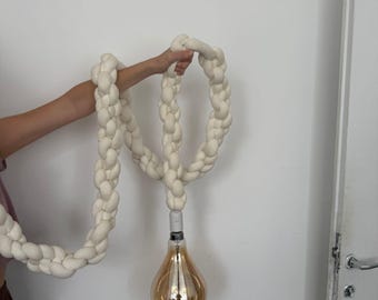 Op de afbeelding: Crèmekleurige, gevlochten touwlamp met een grote, druppelvormige, amberkleurige gloeilamp. Het gevlochten touw wordt door een arm tegen een witte muur gehouden. Een uniek woondecoratie-item.
