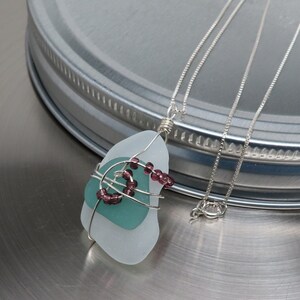 May include: A handcrafted necklace featuring a layered sea glass pendant in shades of blue and teal, wrapped with silver wire. The pendant is adorned with small, dark purple beads and hangs from a delicate silver chain.