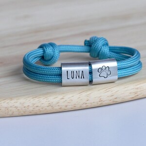 Puede incluir: Un brazalete de paracord azul turquesa con dos dijes plateados. Un dije dice "LUNA" y el otro tiene una huella de pata.