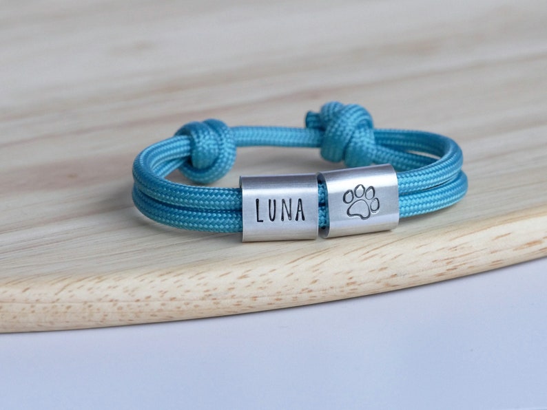Puede incluir: Un brazalete de paracord azul turquesa con dos dijes plateados. Un dije dice "LUNA" y el otro tiene una huella de pata.