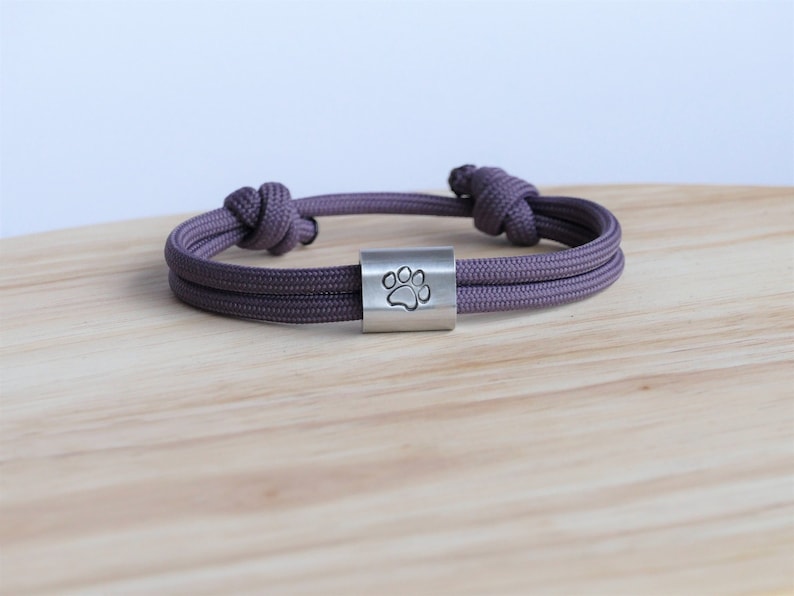 Puede incluir: Una pulsera de paracord morada con un dije plateado con una huella de pata.