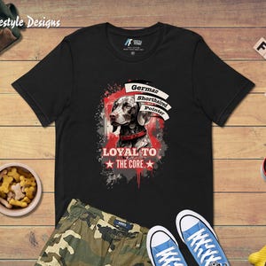 May include: Black t-shirt featuring a graphic of a German Shorthaired Pointer with the text "LOYAL TO THE CORE." The design includes red and white accents. The shirt is displayed with camouflage pants and blue sneakers.