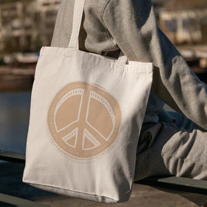 Könnte beinhalten: Eine weiße Canvas-Tragetasche mit einem braunen Peace-Zeichen und dem Text "Spread Love, Give Peace, Be Kind, Stay True,  Love Your Neighbor,  Be The Change,  Spread Joy,  Give Hope,  Be The Light,  Spread Kindness,  Be The Love,  Give Peace,  Spread Love" um das Peace-Zeichen herum.
