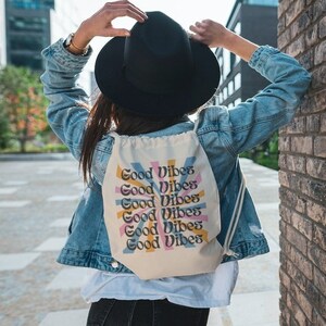 Può includere: Uno zaino bianco in tela con coulisse su cui è ripetuta sei volte la frase "Good Vibes" in un font retrò. Il testo è in uno schema di colori arcobaleno con accenti gialli, rosa e blu. Lo zaino è indossato da una persona che indossa una giacca di jeans e un cappello nero.