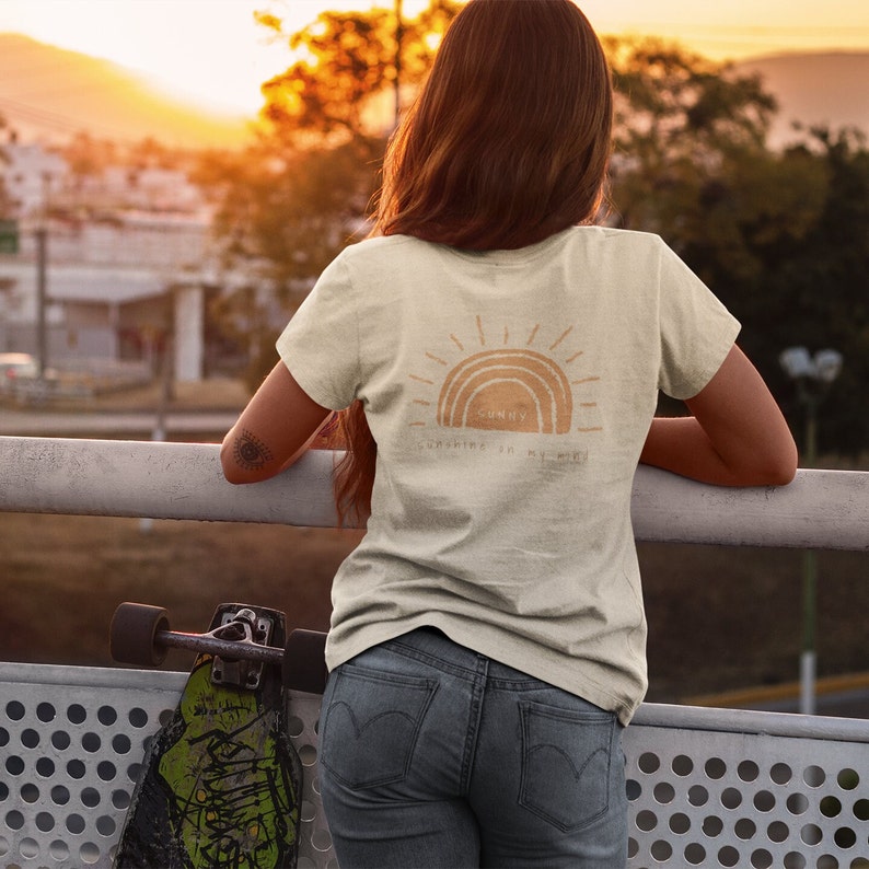 Puede incluir: Una camiseta beige claro con un gr&aacute;fico de un sol y un arco&iacute;ris con el texto "Sunny Sunshine in My Heart".