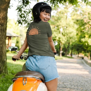 Puede incluir: Una mujer lleva una camiseta verde con un gr&aacute;fico de un sol y el texto "Sunny. Sunshine is in my mind." Ella est&aacute; conduciendo un scooter.