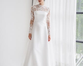 Puede incluir: Un vestido de novia blanco con una superposición de encaje y mangas largas. El vestido tiene un escote alto y una falda fluida.