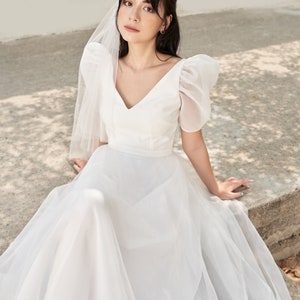 May include: A white wedding dress with a v-neckline and puff sleeves. The dress has a tulle skirt and a sheer veil.