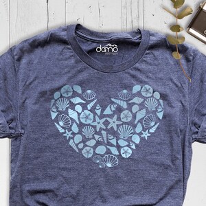 May include: A navy blue t-shirt with a light blue heart-shaped design made of seashells and starfish. The shirt has a crew neck and short sleeves.