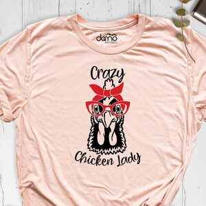 K&ouml;nnte beinhalten: Ein pfirsichfarbenes T-Shirt mit einer Schwarzwei&szlig;illustration eines Huhns, das eine rote Brille und ein rotes Bandana tr&auml;gt. Der Text "Crazy Chicken Lady" ist unter der Illustration aufgedruckt.