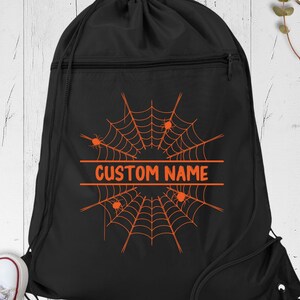 May include: A black drawstring backpack featuring an orange spiderweb graphic with small spiders and the text CUSTOM NAME centered horizontally. The bag has a top zippered compartment and black cord straps for closure and carrying.