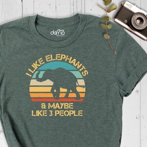 May include: A green t-shirt with a vintage-style graphic of an elephant in front of a sunset. The text on the shirt reads "I like elephants & maybe like 3 people".