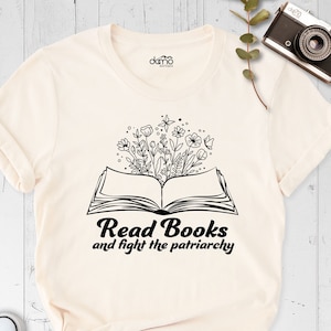 May include: A cream colored t-shirt with a black graphic of an open book with flowers growing out of it. The text "Read Books and fight the patriarchy" is below the book.