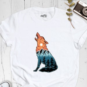 Puede incluir: Camiseta blanca con un gr&aacute;fico de un lobo aullando con un paisaje de monta&ntilde;a dentro de su silueta. El lobo est&aacute; mirando hacia la izquierda y las monta&ntilde;as est&aacute;n en tonos de azul, verde y naranja.