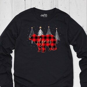 May include: Black long-sleeve shirt featuring a red and black buffalo plaid bear with the name "Agnes" in script. The design includes white trees, gold stars, and snowflakes, creating a festive holiday theme.