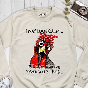 May include: A beige sweatshirt with a graphic of a cartoon chicken wearing red glasses and a red and white polka dot bandana. The text on the sweatshirt reads "I may look calm... But in my head I've pecked you 3 times..."