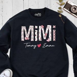 May include: A navy blue sweatshirt featuring the word "MIMI" in a decorative font, with patterns and hearts. Below, the names "Tommy" and "Emma" are written with a heart symbol. The sweatshirt has a crew neck and long sleeves.